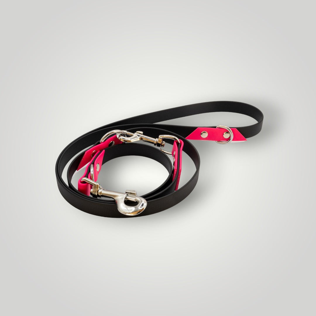 Die Pfotenimglueck Biothane Hundeleine 2-fach verstellbar in Schwarz-Pink mit silbernen Clips liegt kreisförmig auf einem hellgrauen Hintergrund.