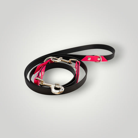 Die Pfotenimglueck Biothane Hundeleine 2-fach verstellbar in Schwarz-Pink mit silbernen Clips liegt kreisförmig auf einem hellgrauen Hintergrund.