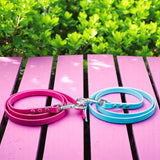 Zwei Pfotenimglueck Biothane® Schleppleinen auf Maß (genietet), ein rosa, ein hellblau, liegen zusammengerollt auf rosafarbenem Holz mit grünen Büschen – ideal fürs Hundetraining dank wasserfestem Material.