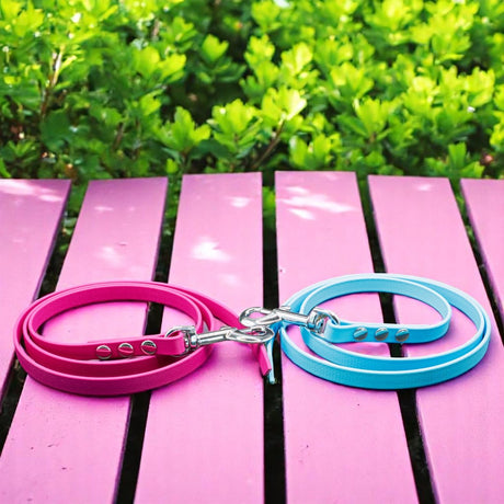 Zwei Pfotenimglueck Biothane® Schleppleinen auf Maß (genietet), ein rosa, ein hellblau, liegen zusammengerollt auf rosafarbenem Holz mit grünen Büschen – ideal fürs Hundetraining dank wasserfestem Material.