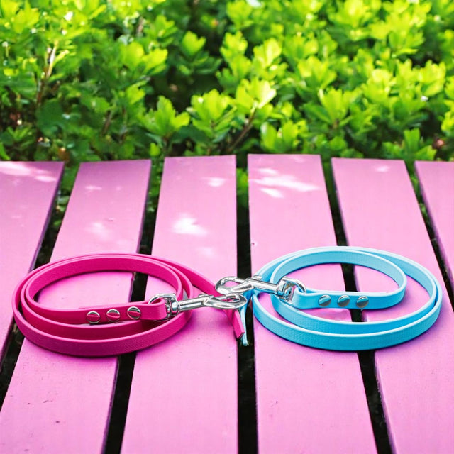 Zwei Pfotenimglueck Biothane® Schleppleinen auf Maß (genietet), ein rosa, ein hellblau, liegen zusammengerollt auf rosafarbenem Holz mit grünen Büschen – ideal fürs Hundetraining dank wasserfestem Material.