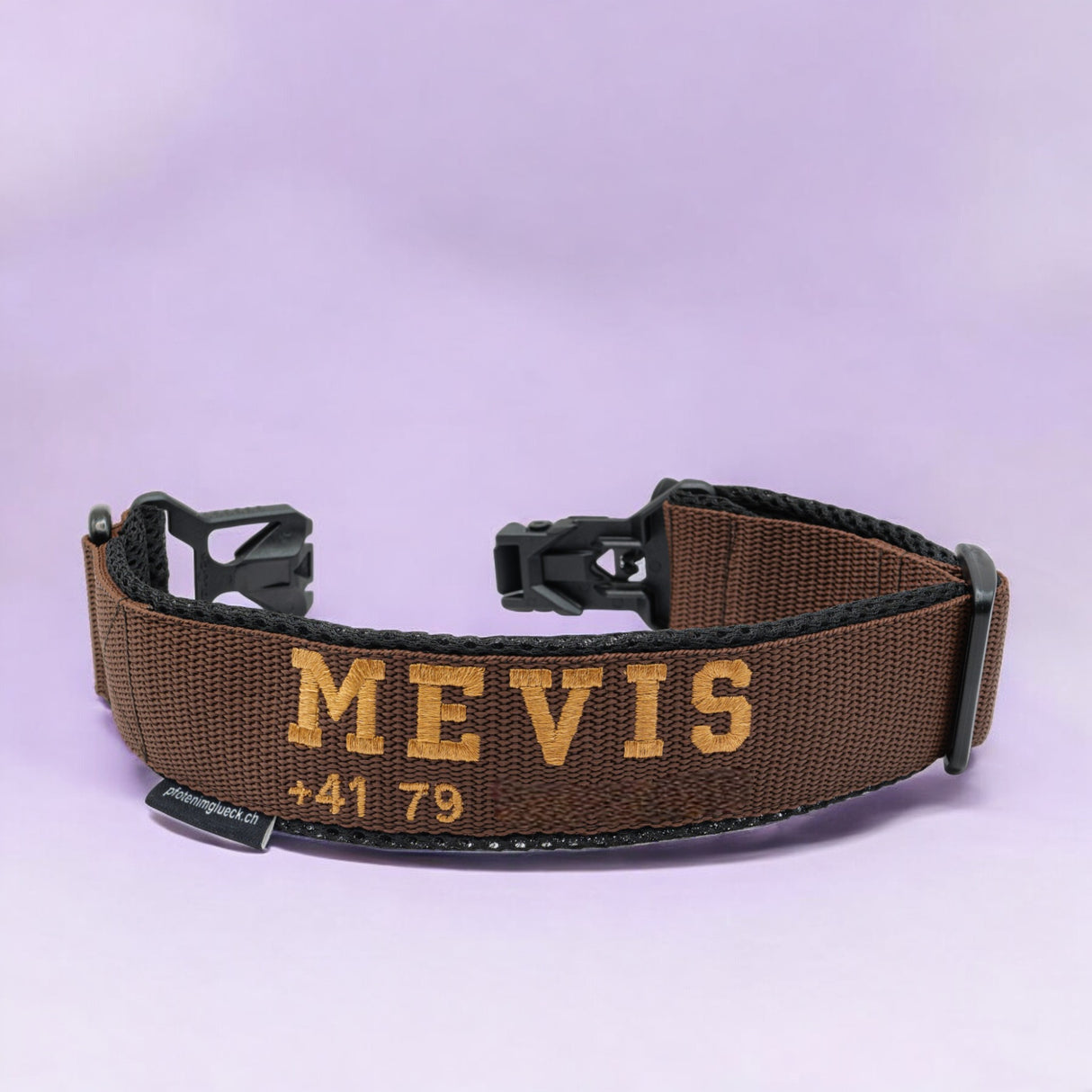 Ein personalisiertes, braunes besticktes Hundehalsband von pfotenimglück mit einem FIDLOCK® Magnetverschluss und schwarzen Kunststoffschnallen, bestickt mit dem Namen „MEVIS“ und einem Teil der Telefonnummer „+41 79“, steht auf einem hellvioletten Hintergrund.