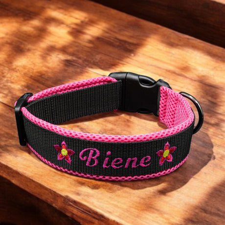 Ein schwarz-rosa „Besticktes Hundehalsband Steckverschluss 'Selbstgestalten'“ von pfotenimglück liegt auf Holz, bestickt mit „Biene“ und Blumenmotiven. Dieses personalisierte Halsband ist robust konstruiert und verfügt über eine Schnalle und einen Metallring zur Befestigung der Leine.