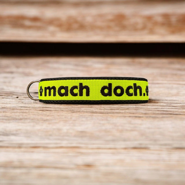 Ein gelber Stoff-Schlüsselanhänger mit schwarzen Rändern und „mach doch.“ in kräftigen schwarzen Buchstaben auf einer Holzoberfläche – perfekte Inspiration für ein pfotenimglück Hundehalsband mit Zugstopp, personalisierbar und mit Bestickung.