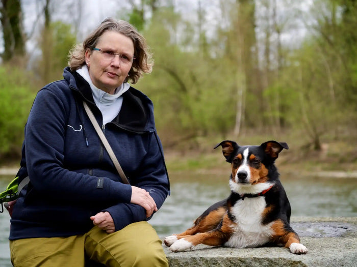 Eine Person mit kurzen Haaren, Brille und dunkler Jacke sitzt auf einem Felsvorsprung an einem Gewässer. Ein schwarz-brauner Hund liegt daneben. Im Hintergrund sind Bäume zu sehen.
