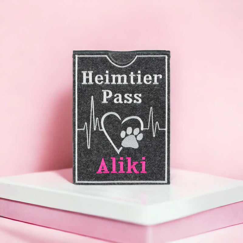 Ein grauer Kartenhalter mit den Worten „Heimtier Pass“ und „Aliki“ in weißer und rosa Schrift, mit einem Herzschlaglinien-, Herz- und Pfotendesign. Er liegt auf einer weißen und rosa Oberfläche vor einem rosa Hintergrund.