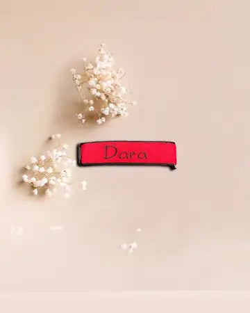 Auf einem beigen Untergrund ist ein roter Stoffstreifen mit der schwarzen Aufschrift „Dara“ platziert, umgeben von kleinen, zarten weißen Blüten.