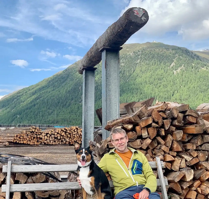 Ein Mann in einer grünen Jacke sitzt auf einer Holzbank, neben ihm ein schwarz-weißer Hund. Dahinter sind gestapelte Baumstämme und eine hohe Holzkonstruktion zu sehen. Im Hintergrund ist ein grüner Berg unter einem blauen Himmel mit Wolken zu sehen.