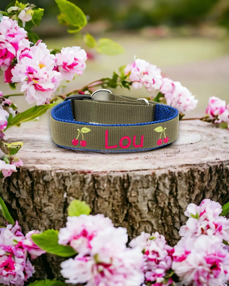 Ein Hundehalsband mit dem Namen „Lou“ und Kirschdekorationen liegt auf einem Baumstumpf, umgeben von blühenden rosa und weißen Blumen.