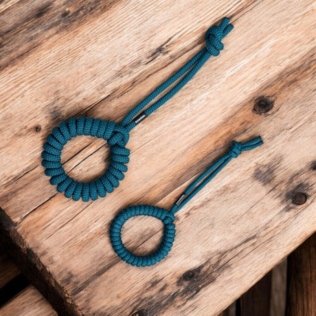 Auf einer rustikalen Holzoberfläche liegen zwei blaue Paracord-Schlüsselanhänger mit kreisförmigen Mustern und geschlungenen Enden, die einen Kontrast zur natürlichen Textur des Holzes bilden und den verspielten Charme von pfotenimgluecks „treusinn“ widerspiegeln. Tau-Spielzeug LOOPY Petrol.