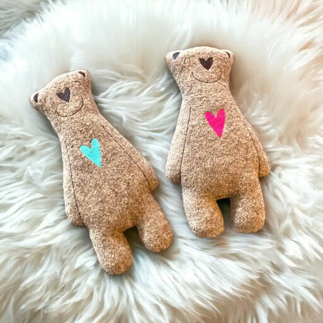 Zwei Plüschteddybären liegen auf einer flauschigen weißen Fläche. Der linke Bär hat ein blaues Herz auf der Brust, der rechte ein rosa Herz. Beide haben schlichte, gestickte Gesichtszüge.