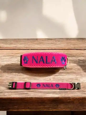 Zwei Hundehalsbänder mit dem Namen „NALA“ und Pfotenabdruck in Blau. Die Halsbänder sind rosa und liegen auf einer Holzoberfläche. Ein Halsband steht aufrecht, das andere liegt flach.